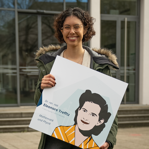Porträtaufnahme einer Frau, die ein Schild in die Kamera hält, das ein gezeichnetes Porträt von Eleonore Trefftz zeigt.