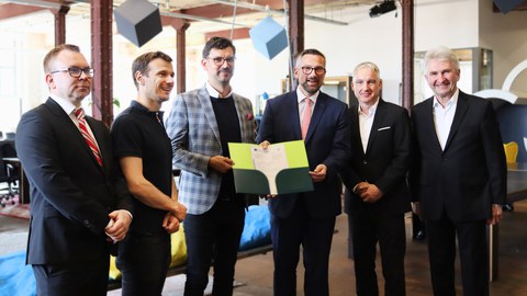 Gruppenbild mit 6 Herren nebeneinander im Anzug. Die zwei Herren in der MIttel halten gemeinsam eine Urkunde in einer Mappe in den Händen. Alle Herren schauen freundlich.