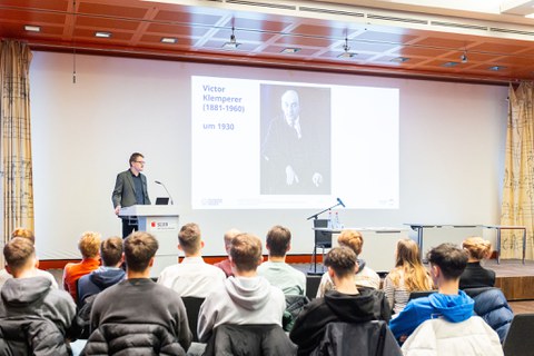 Blick in einen Vortragssaal. Ein Mann steht an einem Rednerpult. Im Vordergrund sieht man Schüler:innen, die zuhören.