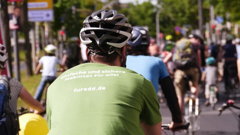 Foto von Kopf und Rücken eines Fahrradfahrers. Er hat einen grau-weißen Fahrradhelm und ein hellgründes T-Shirt mit der weißen Aufschrift: Einfache und sichere, darunter: Mobilität für alle! Darunter: furedde.de. Vor ihm sind verschwommen andere Fahrradfahrer von hinten zu sehen.