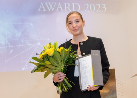 Junge Frau mit blonden, zusammengebundenen Haaren steht mit einer Urkunde in der einen und einem Blumenstrauß in der anderen Hand auf einer Bühne.