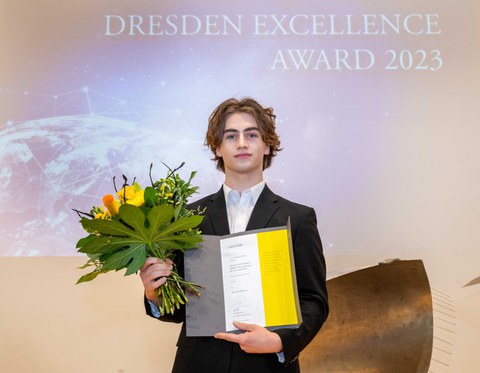 Junger Mann mit dunklen lockigen Haaren steht mit einer Urkunde in der einen und einem Blumenstrauß in der anderen Hand auf einer Bühne.