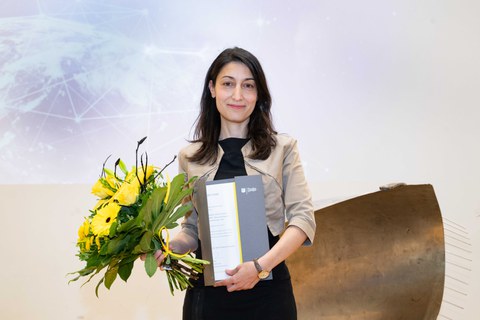Junge Frau mit langen dunklen Haaren steht mit einer Urkunde in der einen und einem Blumenstrauß in der anderen Hand auf einer Bühne.