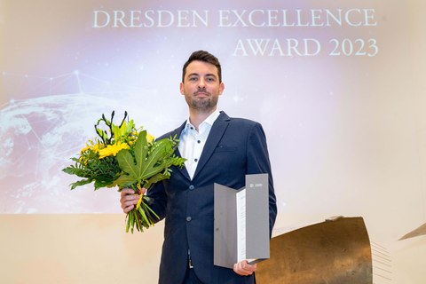 Junger Mann mit kurzen dunklen Haaren steht mit einer Urkunde in der einen und einem Blumenstrauß in der anderen Hand auf einer Bühne.