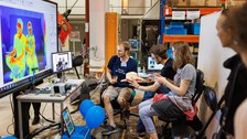 Foto von zwei Personen, die auf Stühlen sitzen. Vor ihnen befindet sich ein Monitor, der sie im Wärmebild zeigt.