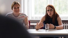 zwei Frauen an einem Tisch. Eine redet, die andere lächelt.
