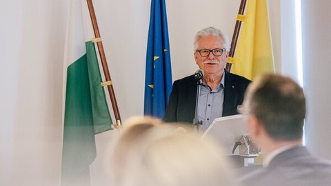 Foto eines Herrn mit weißen Haaren und Schnauzbart und Brille vor drei Flaggen stehend. v.l. weiß-grün, blau, gelb. Im Vordergrund sind die verschwommenen Hinterköpfe zweier Personen zu sehen.