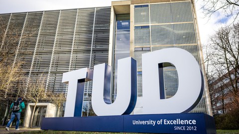 Photograph of a large sculpture of three large white 3D letters "TUD" on a blue pedestal. The sculpture is located on a lawn with the Auditorium Center in the background.