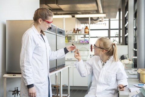 Photo of two women in the laboratory. One shows the other the result in the test tube.