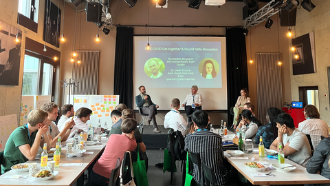Fotoaufnahme von drei Personen, die auf einer Bühne an einer Podiumsdiskussion teilnehmen. Vor der Bühne befindet sich eine Gruppe von Zuhörerinnen und Zuhörern, die an Tischen sitzen.