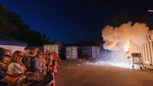 Foto einer Puppe, bei der gerade etwas explodiert und in Flammen steht. Aus sicherem Abstand wird die Szene von Zuschauenden beobachtet.