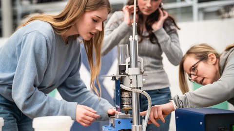 Fotoaufnahme von drei jungen Frauen, die an einer Werkbank stehen, auf dem sich eine Presse befindet.