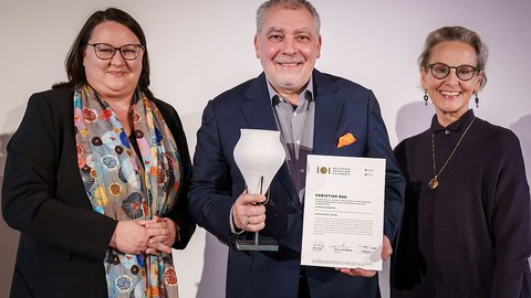 Katrin Stump (links) und Prof.in Ursula Staudinger (rechts) übergeben die Urkunde und Gefäßstatuette an Spitzenkoch Christian Bau.