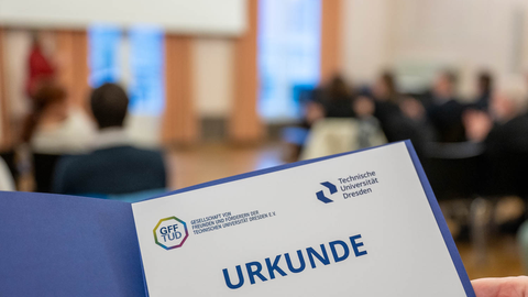 Foto von einer Urkunde auf der das Logo der GFF und der TU Dresden abgebildet sind. Im Hintergrund sind unscharf Menschen zu erkennen, die an einer Veranstaltung teilnehmen.