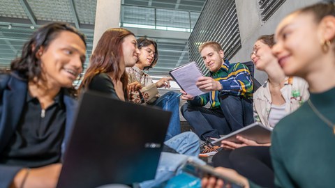 Foto von sechs Studierenden, die auf dem TUD-Campus auf einer Treppe sitzen und zusammen lernen. 