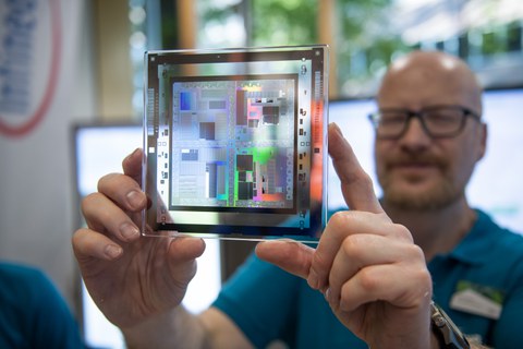 Eine Person hält eine transparente quadratische Platte direkt vor die Kamera. Darauf befindet sich eine bunte, reflektierende Mikrochip‑ oder Schaltkreisstruktur. Die Person dahinter ist unscharf zu sehen.