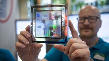 Eine Person hält eine transparente quadratische Platte direkt vor die Kamera. Darauf befindet sich eine bunte, reflektierende Mikrochip‑ oder Schaltkreisstruktur. Die Person dahinter ist unscharf zu sehen.