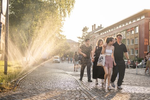 Eine Gruppe junger Menschen läuft lachend eine Kopfsteinpflasterstraße entlang. Links sprühen Rasensprenger feinen Wassernebel in die warme Abendsonne, wodurch die Umgebung golden schimmert.