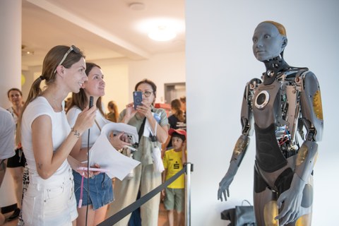 Mehrere Menschen stehen vor einem lebensgroßen humanoiden Roboter in einem Ausstellungsraum. Zwei Personen sprechen gerade in ein Mikrofon, andere fotografieren oder beobachten den Roboter aufmerksam.