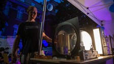 Eine Person steht abends unter einem Zelt und erklärt eine beleuchtete physikalische Apparatur mit runden Glas- oder Kunststoffscheiben. Die Szene ist in blau-lila Licht getaucht, und im Hintergrund sitzen Menschen an Tischen.