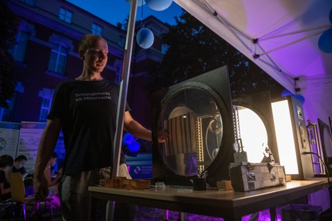 Eine Person steht abends unter einem Zelt und erklärt eine beleuchtete physikalische Apparatur mit runden Glas- oder Kunststoffscheiben. Die Szene ist in blau-lila Licht getaucht, und im Hintergrund sitzen Menschen an Tischen.