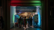 Ein Eingang ist bunt beleuchtet und mit einem Banner „Dresdner Lange Nacht der Wissenschaften“ versehen. Menschen gehen hindurch zu einer Ausstellung mit technischen Geräten und Informationspostern im Inneren.