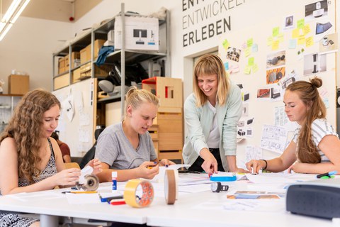 Drei junge Frauen sitzen an einem Tisch und basteln, eine Frau steht dazwischen und zeigt auf die Bastelei.