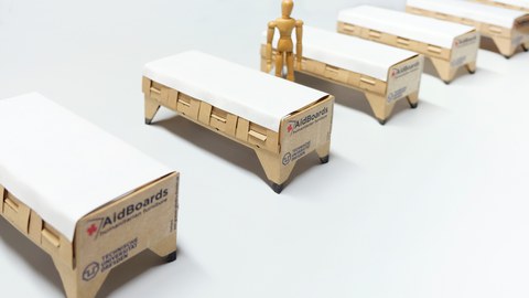 Auf dem Foto sind fünf, nebeneinander schräg von links unten nach rechts oben aufgestellte, Feldbetten aus Pappe zu sehen, neben dem 2. Bett von links steht eine Puppe.