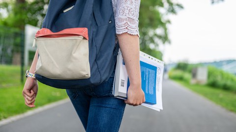 Eine Studentin spaziert am Blauen Wunder Dresden. Sie trägt einen blauen Rucksack auf dem Rücken und hält Hefter und Buch in der rechen Hand.