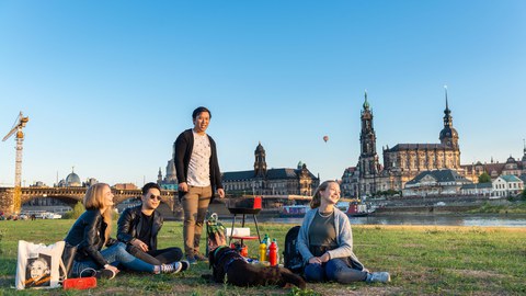 Eine Gruppe junger Menschen sitzt auf der Elbwiese, im Hintergrund die Dresdner Kulisse mit Hofkirche.