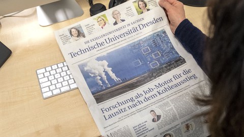Vor einer hellen Tischplatte hält eine Person eine Zeitungsseite ins Bild.