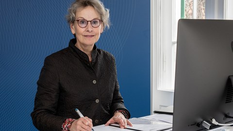 Frau Professorin Staudinger sitzt vor einer blauen Wand, vor sich eine aufgeschlagene Mappe und einen Laptop.