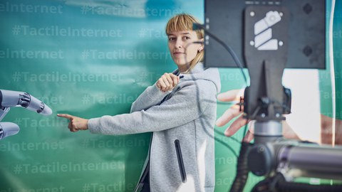 eine junge Frau steht vor einer Wand in blau-grün mit dem mehrfachen Schriftzug #TactileInternet steht und zeigt mit dem Zeigefinger nach vorn.