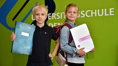 Ein Junge und ein Mädchen stehen mit ihren Zeugnissen vor einer grünen Wand.