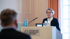 Eine Frau spricht an einem Rednerpult zu Publikum.