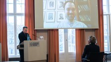 Ein Mann an einem Rednerpult spricht zu einem Mann, der mit Hilfe einer Videokonferenz zugeschaltet ist.