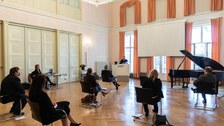 Personen in einem Saal verfolgen die Rede eines Mannes.