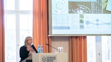 Eine Frau steht an einem Rednerpult und hält einen Vortrag. Im Hintergrund ist eine Prsäentation zu erkennen.