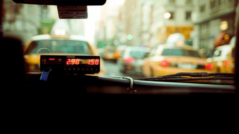 Foto des Innenraums eines Taxis mit Taxometer, man blickt durch die Frontscheibe auf eine start befahrene Straße