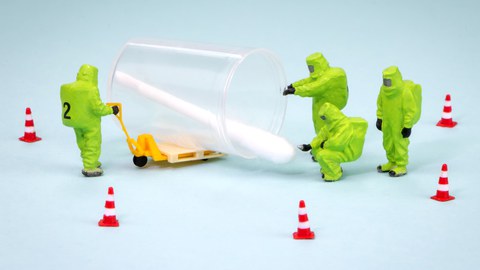 Vier Figuren im grünen Schutzanzug umringen ein übermannsgroßes Wattestäbchen, ringsherum sind Verkehrspylonen aufgestellt.