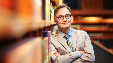 Prof. Bonte lehnt mit verschränkten Armen an einem mit Büchern gefüllten Regal, im Hintergrund sieht man verschwommen Bücherregale.