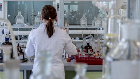 Man sieht eine junge Frau mit Zopf von hinten, die inmitten von chemischen Gerätschaften in einem Labor steht.
