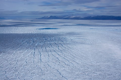 Eine große, rissige Eisfläche, im Hintergrund sind Berge und der Himmel zu sehen, alles in verschiedenen Schattierungen von Hellblau.