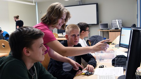 Schüler sitzen nebeneinander an einer Schulbank und sehen auf Bildschirme. Ein Mädchen steht zwischen ihnen und zeigt auf einen Bildschirm.