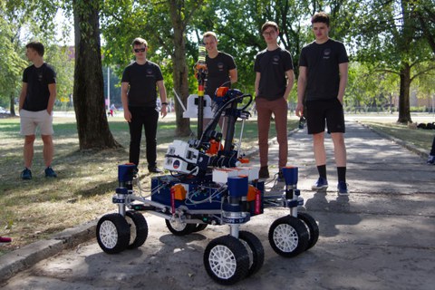 Mars-Rover auf einem Weg, dahinter fünf junge Männer.