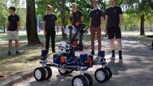 Mars-Rover auf einem Weg, dahinter fünf junge Männer.