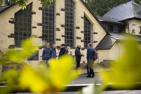 Personengruppe vor einem hellen, schiefergedeckten Gebäude.