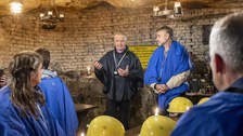 Personen in Schutzkleidung sitzen an hölzernen Tischen in einem unterirdischen Raum. Vorn stehen zwei redende Männer.