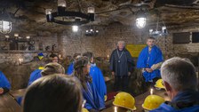 Personen in Schutzkleidung sitzen an hölzernen Tischen in einem unterirdischen Raum. Vorn stehen zwei redende Männer.