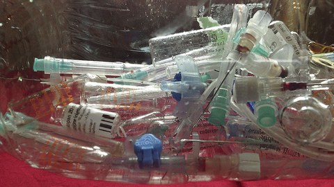 Abfall von gebrauchten Spritzen und kleine Behältnisse liegen in durchsichtigem Plastikbehälter.